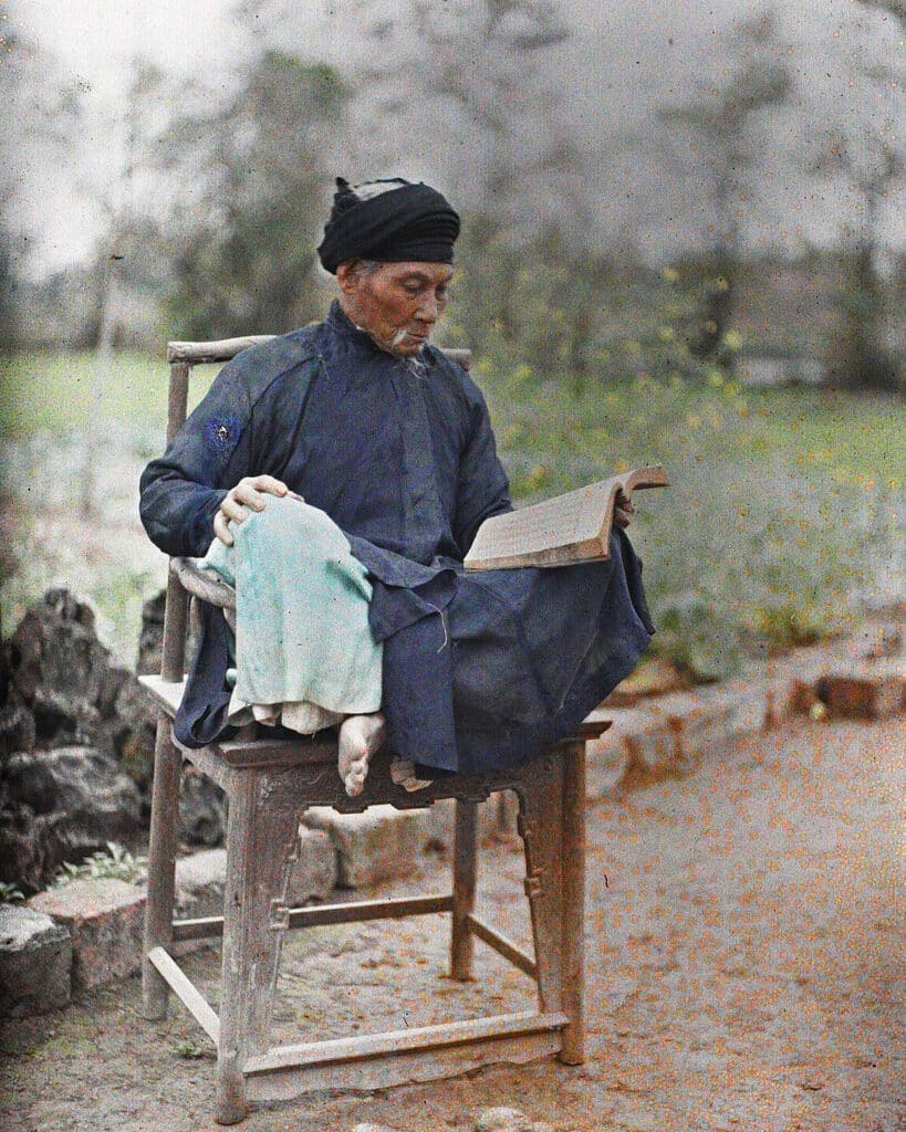 Ảnh màu năm 1915: một cụ đồ áo the, khăn xếp, ngồi trên ghế gỗ trong vườn ở Hà Nội, chăm chú đọc sách